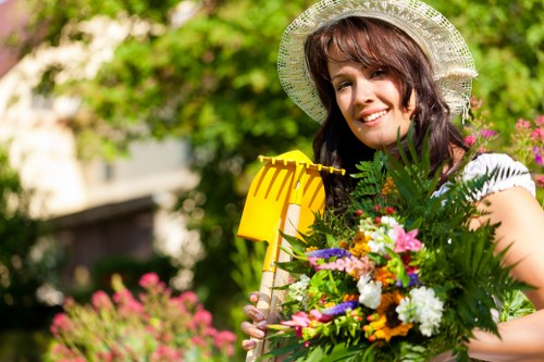 A woman stands in a well-maintained garden with a variety of plants and vibrant flowers, holding a yellow gardening tool and a large bouquet of mixed blooms and greenery. Behind her, the garden features a lush, green lawn with neatly trimmed edges and a paved patio area made of textured stone slabs. In the background, mature shrubs and small trees with dense foliage provide natural borders, while colorful flower beds border the lawn with a mix of pinks, purples, whites, and yellows. The scene is bathed in natural daylight, suggesting a bright, fair-weather day, with soft shadows indicating gentle sunlight filtering through the garden’s leafy canopy. The overall setting appears tidy and inviting, characteristic of professional garden maintenance, possibly in the Palmers Green area of London, highlighting landscaping and gardening services available to enhance outdoor spaces in the local community.